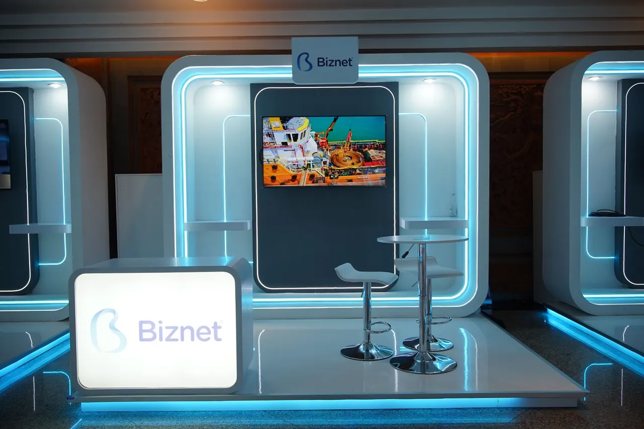 Biznet booth with a lit logo counter, wall screen and high stools, outlined in cyan light
